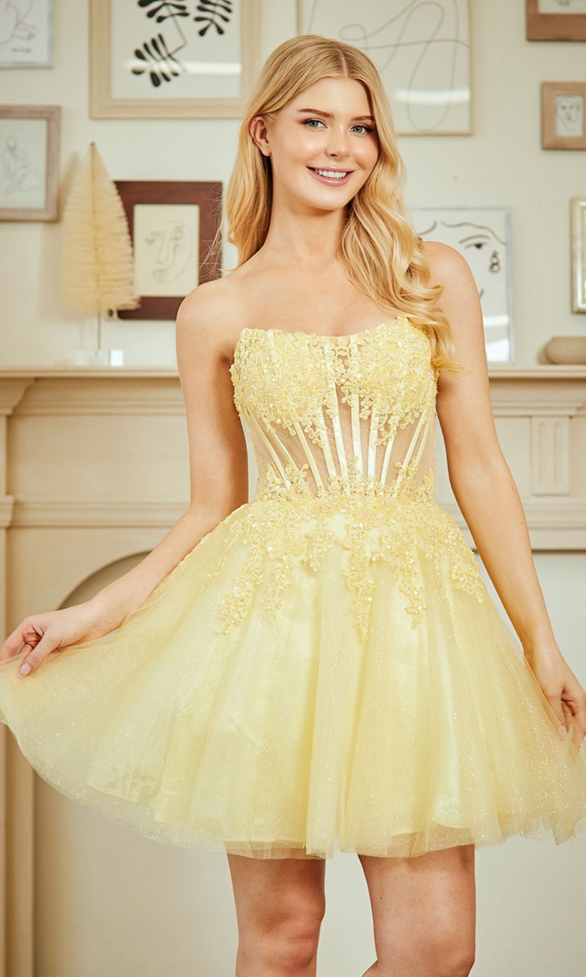 A woman stands indoors holding the sides of her yellow Amelia Couture 9083S strapless short party dress, which features a sheer bodice and floral appliqué.